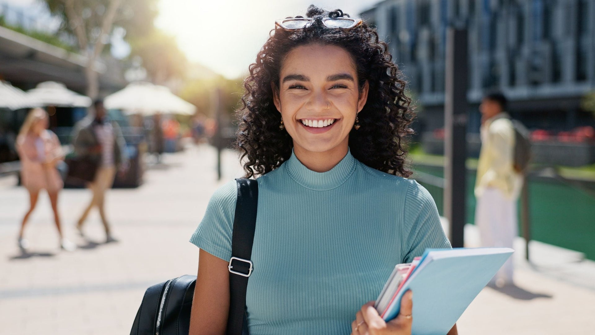 A young woman with curly hair smiling happily while holding books or notebooks, outdoors on a sunny day, with blurred pedestrians in the background.