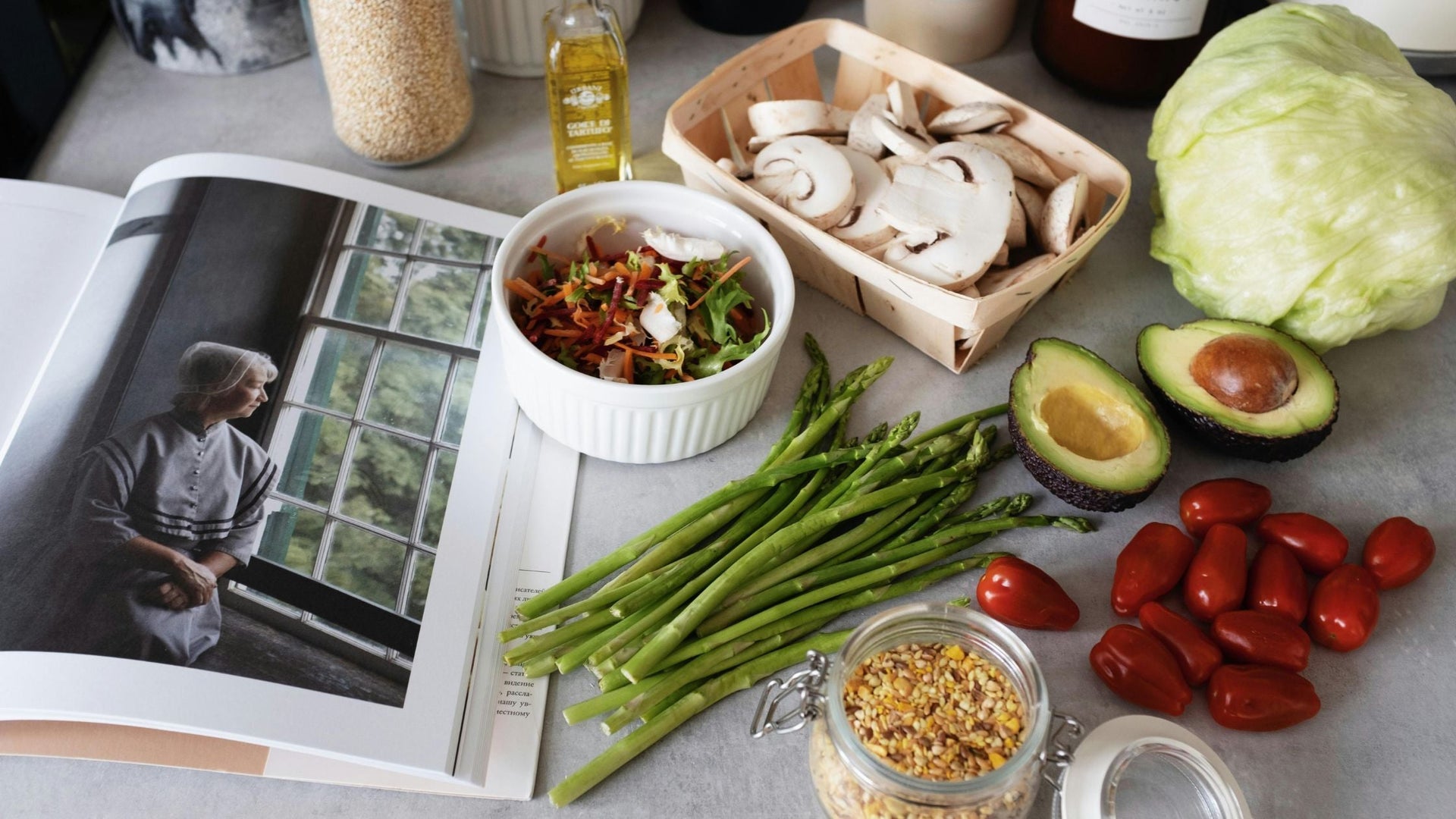 This image depicts a variety of fresh ingredients laid out for cooking, including asparagus, cherry tomatoes, sliced mushrooms, avocado, and a head of lettuce. There's also a bowl with a salad mix, a jar of grains or seeds, and a bottle of olive oil.