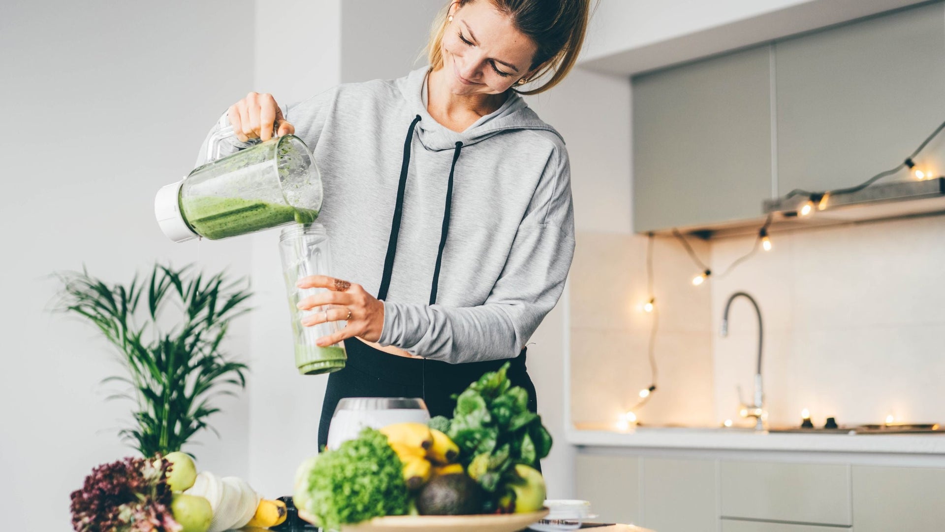 In this image, a woman is pouring a green smoothie or juice into a glass in a modern kitchen. The countertop is adorned with various fresh fruits and vegetables like bananas, avocados, and leafy greens, suggesting a focus on healthy living and nutrition. 