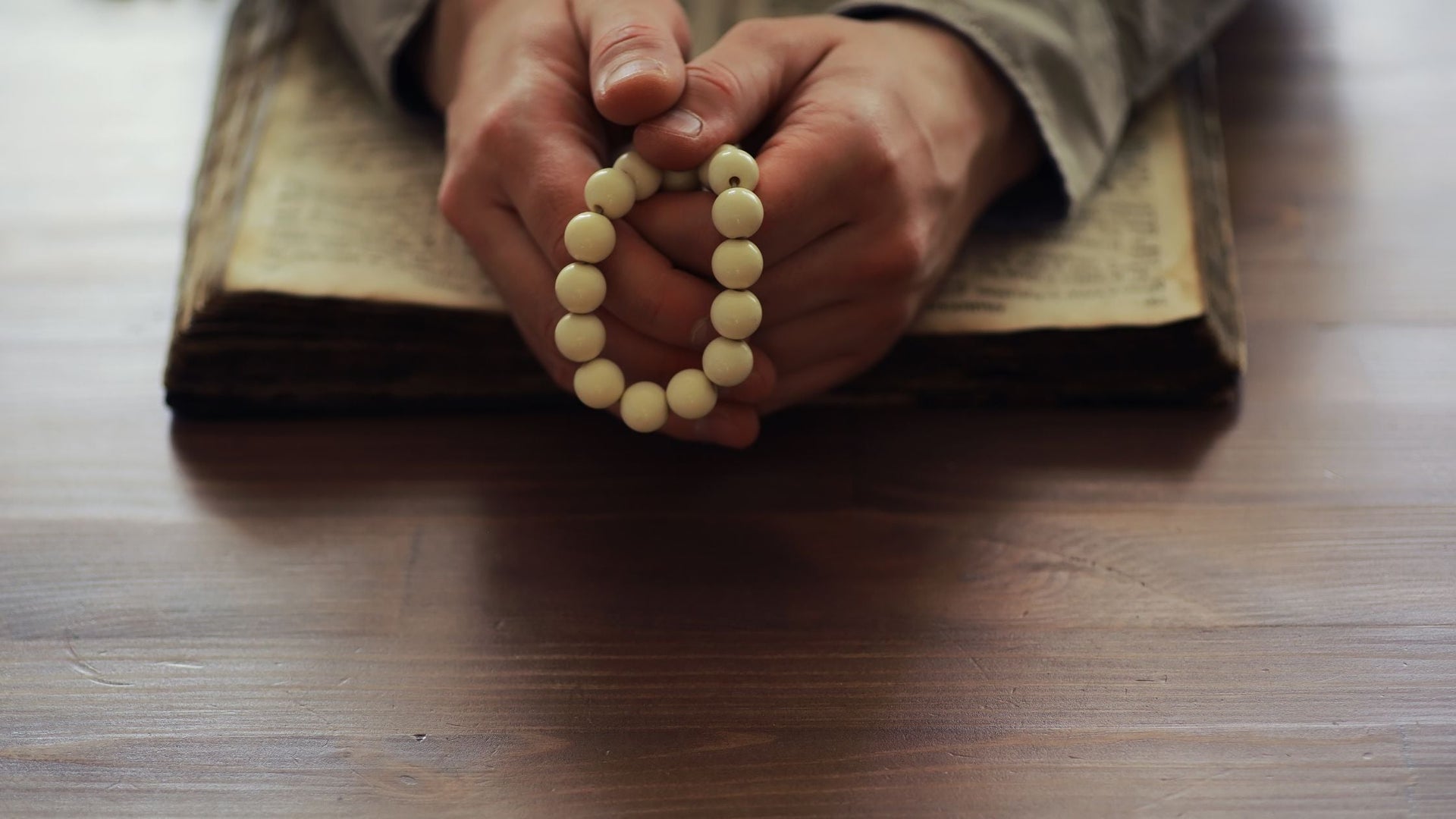 This image features a person holding a prayer bead or rosary, with their hands resting on an open religious book, likely a Bible or similar sacred text. The serene and contemplative setting emphasizes prayer, devotion, and spiritual reflection.